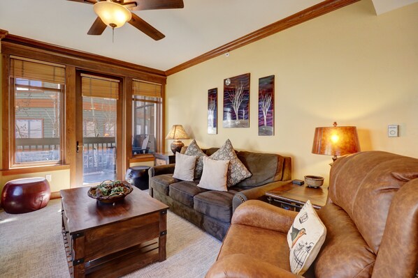 A living room with a brown leather sofa, a wooden coffee table, and a lamp. There are three paintings on the wall and a ceiling fan. - Mc204 Mill Club 3 Bedroom Condo (Copper Mountain)