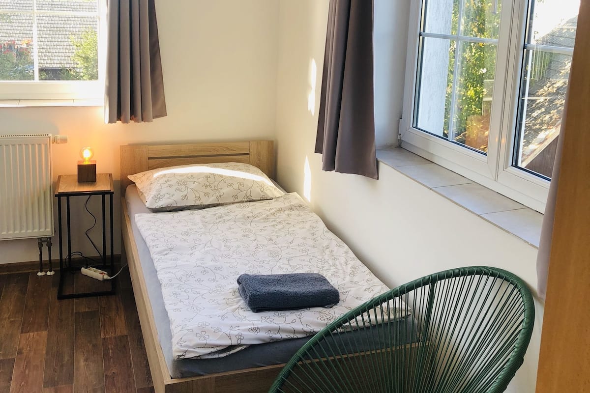 A small, neatly arranged bedroom with a single bed, a bedside table with a lamp, a chair, and a window with curtains.