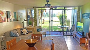 A spacious living room with a wooden dining table, a sofa, and a large window offering a view of a garden.