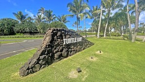 A stone wall with "Turtle Bay" inscribed on it, surrounded by lush green grass and palm trees under a clear blue sky.