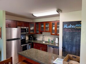 A compact kitchen with a stainless steel refrigerator, microwave, and stove, wooden cabinets, a granite countertop, and a chalkboard with a welcome message.