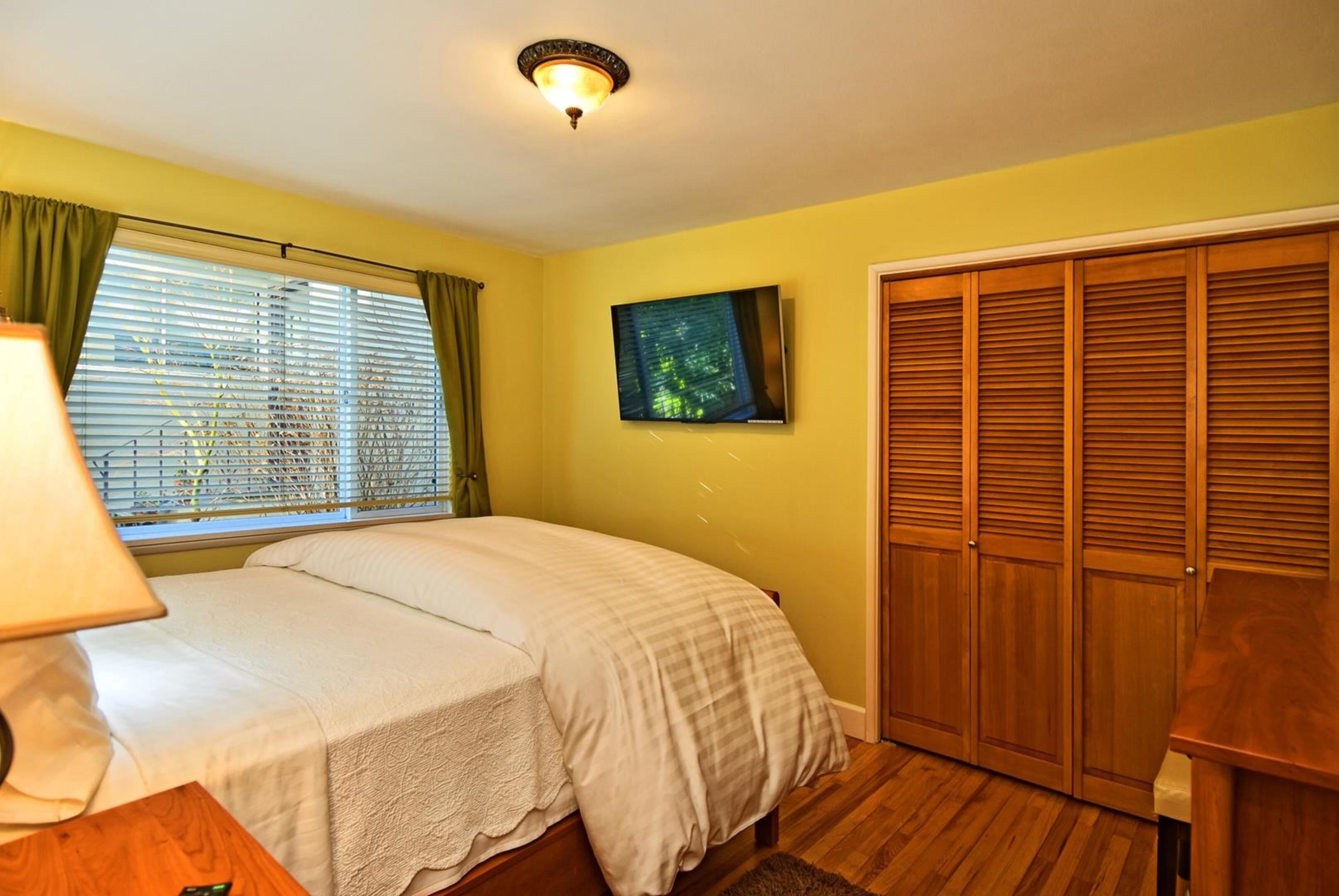 A bedroom with a bed, a TV on the wall, a window with blinds, and a wooden wardrobe.