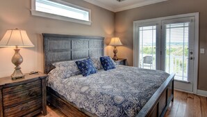 Une chambre à coucher avec un grand lit, deux tables de chevet, deux lampes et une vue sur l’extérieur, visible à travers une porte.