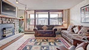 Ein gemütliches Wohnzimmer mit Kamin, einem braunen Ledersofa, einem gemusterten Teppich und einem großen Fenster mit Blick auf Gebäude.