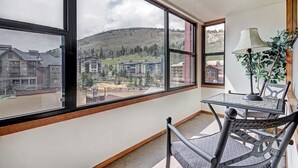 Ein Zimmer mit großen Fenstern, das einen Blick auf eine gebirgige Landschaft und Gebäude bietet. Ein Tisch mit einer Lampe und ein Stuhl sind ebenfalls vorhanden.
