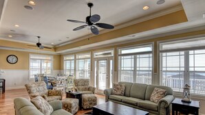 A spacious living room with a large sofa set, a coffee table, and a ceiling fan. There are multiple windows providing natural light and a view of the city. - Luxury 5BR · Sleeps 17 · 1st St · 50 Yards to Beach · Pool · Elevator (Ocean Isle Beach)