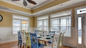 A dining room with a wooden table set for six, surrounded by colorful chairs, large windows with blinds, and a ceiling fan. - Luxury 5BR · Sleeps 17 · 1st St · 50 Yards to Beach · Pool · Elevator (Ocean Isle Beach)