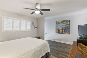A bedroom with a bed, a ceiling fan, a painting, a television, and a small table.