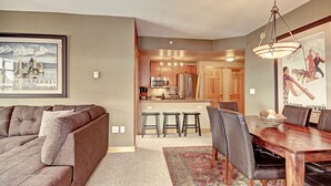 A living room with a sofa, a dining table with chairs, a kitchen area with a refrigerator and microwave, and a hallway leading to a door.
