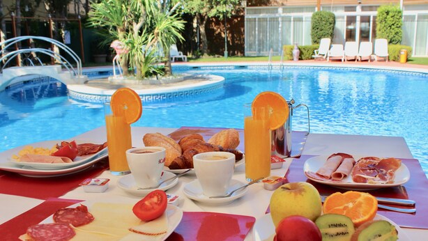 A breakfast table with croissants, pastries, fruit, and coffee by a pool. - Hotel Ampuria Inn (Castello d'Empuries)