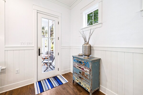 A hallway with a white door, a blue and white striped rug, a vintage blue cabinet with decorative items, and a window with a view of greenery. - 'Seas the Day' in WaterColor Beach 5 Bdrm/Slps 10| Steps to Dragonfly Pool! (Santa Rosa Beach)