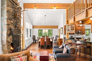A spacious living area with a stone fireplace, wooden beams, a dining table with chairs, and a kitchen with wooden cabinets and a bar area. - RMR: The Village House in Teton Village (Teton Village)