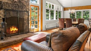 A stone fireplace with a fire, a wooden ceiling, a dining area with chairs, and a large window. - RMR: The Village House in Teton Village (Teton Village)