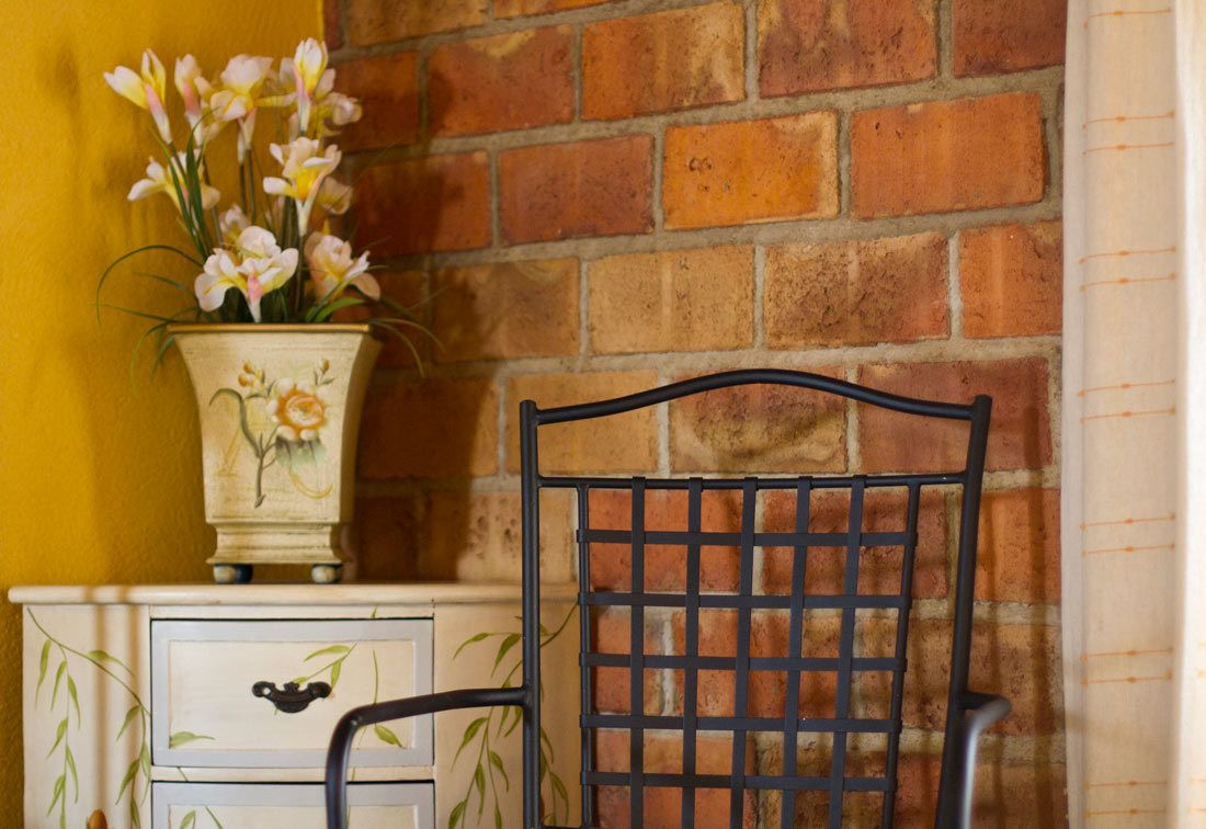 A chair with a metal grid backrest against a brick wall, a vase with flowers on a dresser, and a yellow wall.