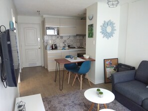 Un espacio habitable compacto con kitchenette, una zona de comedor con sillas azules, un sofá gris y una pequeña mesa blanca con plantas.