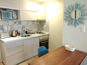Una cocina compacta equipada con microondas, hervidor y estufa a gas. Se observa un lavamanos, una mesa de madera y una silla azul. La cocina presenta baldosas con diseño y un espejo decorativo en la pared.