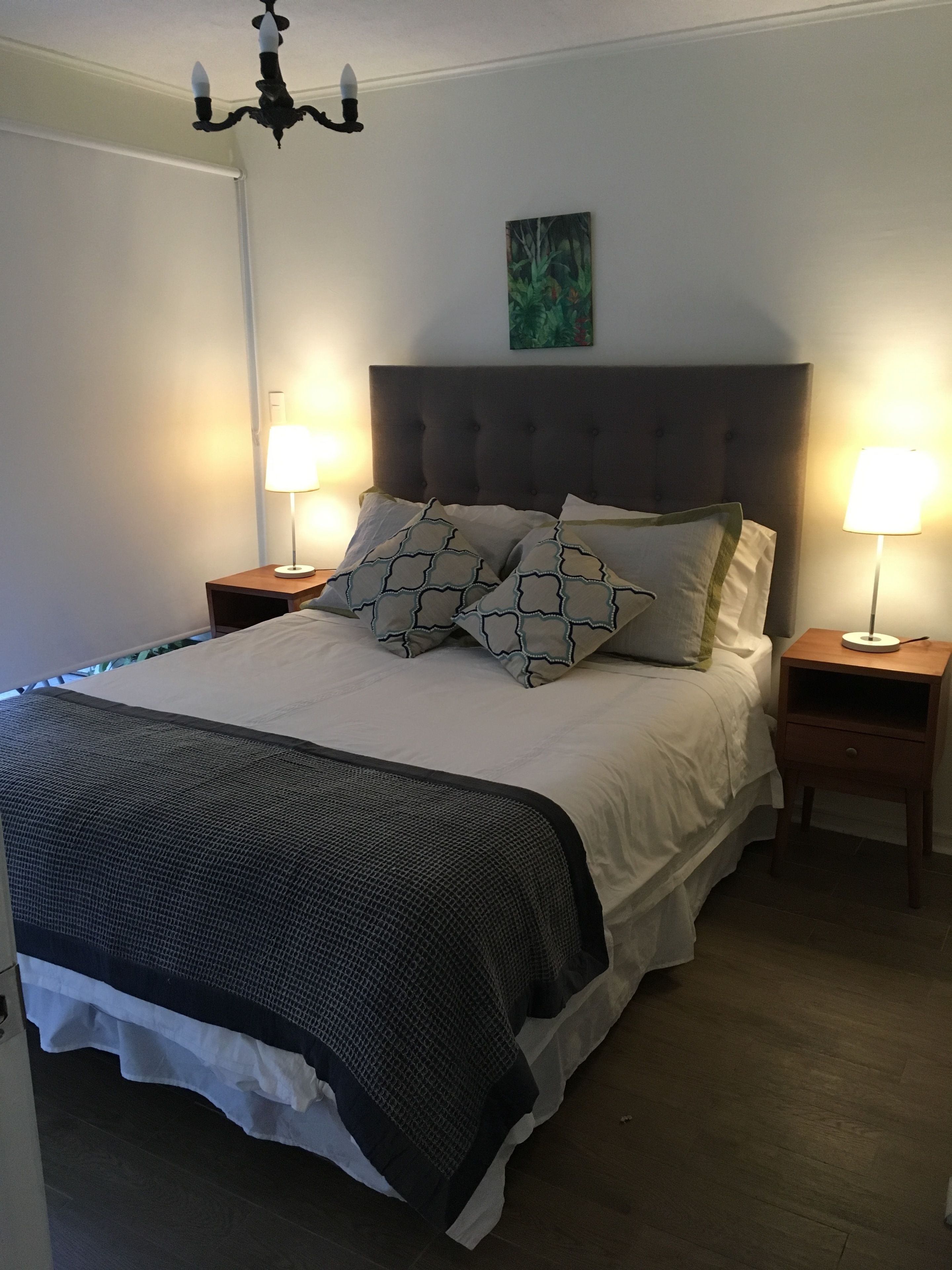 Ein ordentlich bezogenes Bett mit grauem Kopfteil, zwei Nachttische mit Lampen und ein Bild an der Wand.