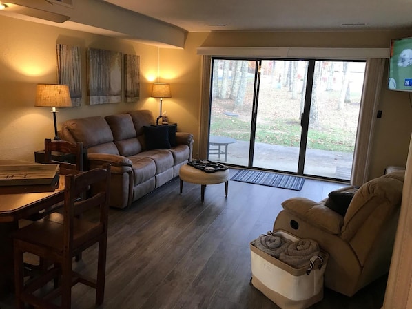 A cozy living room with a brown leather sofa, a wooden coffee table, and a beige armchair. There is a sliding glass door leading to a patio area with a view of trees and a lawn.