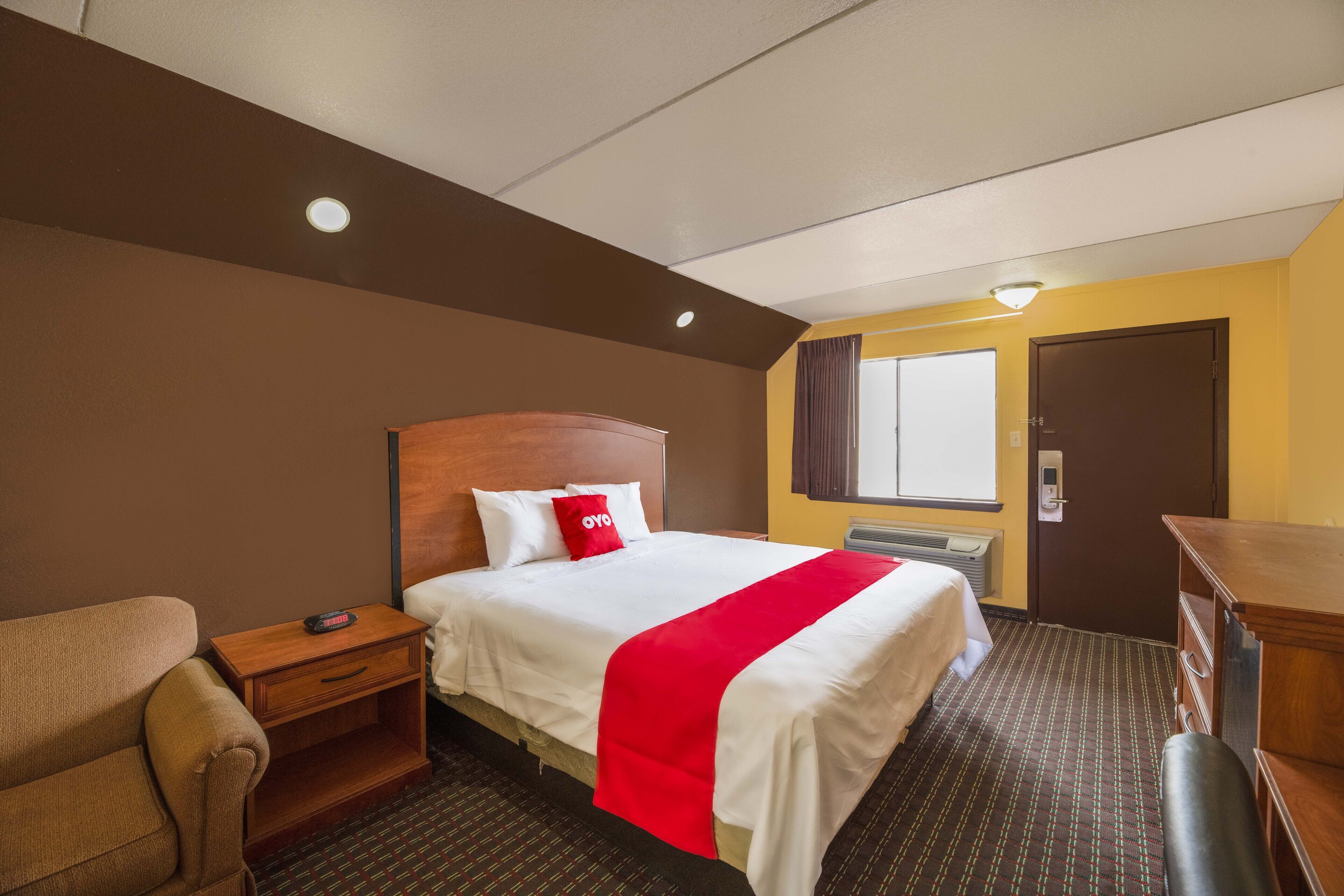 A hotel room with a bed, a wooden nightstand, a desk, a chair, and a window with curtains.
