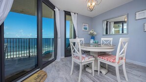 Un comedor con vistas al océano, una mesa redonda, sillas blancas con cojines estampados y un jarrón azul con flores.