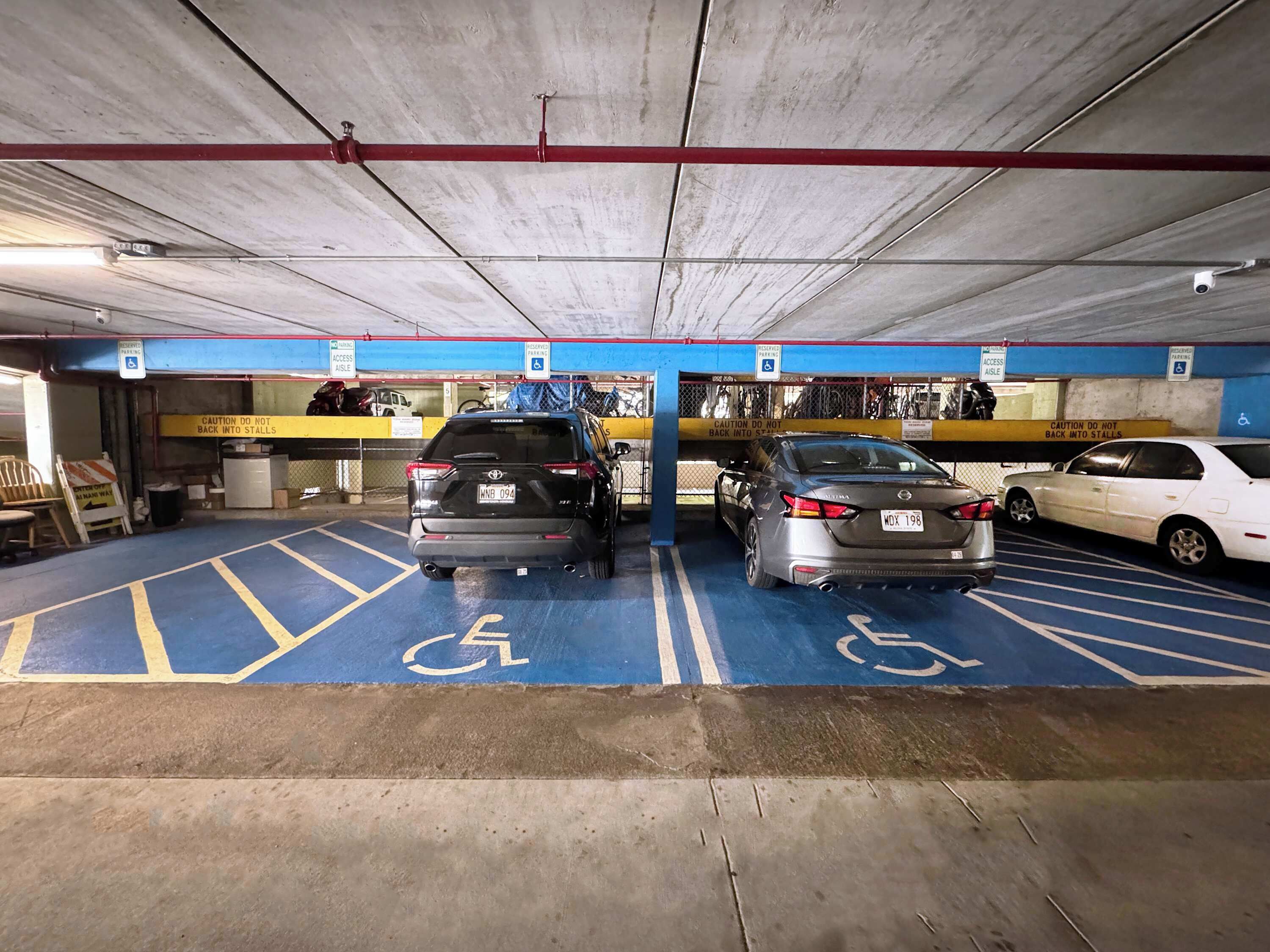 A parking garage with blue handicapped parking spaces, a red pipe on the ceiling, and a sign warning not to back into stalls.