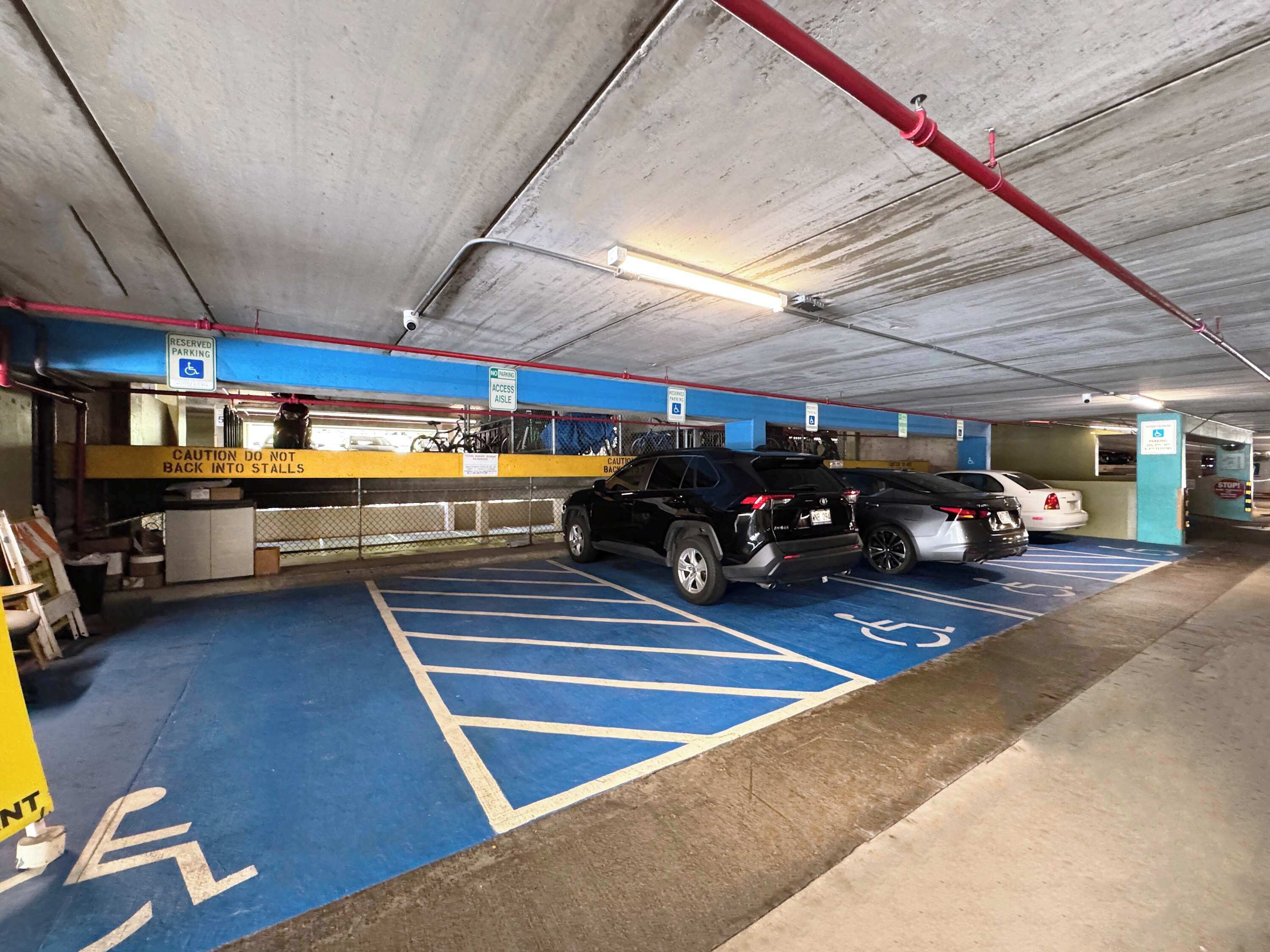 A parking garage with blue handicapped parking spaces, a yellow caution sign, and a red pipe on the ceiling.