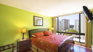 Un dormitorio con cama, mesitas de noche, una ventana con vista a edificios y un televisor de pared.