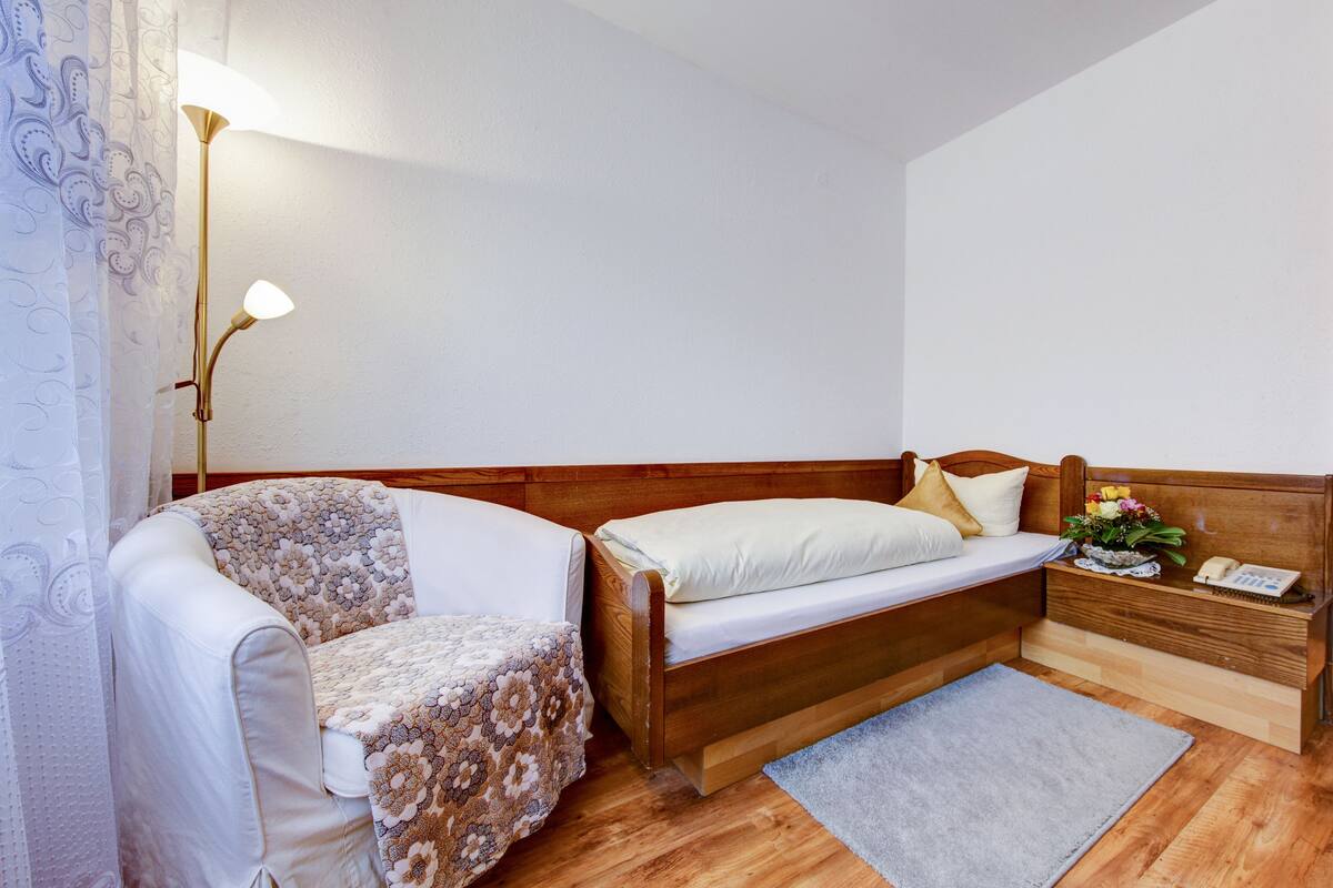 Ein kleiner, gemütlicher Raum mit einem Einzelbett, einem Sessel, einem Nachttisch mit Lampe und einem kleinen Holzbeistelltisch.