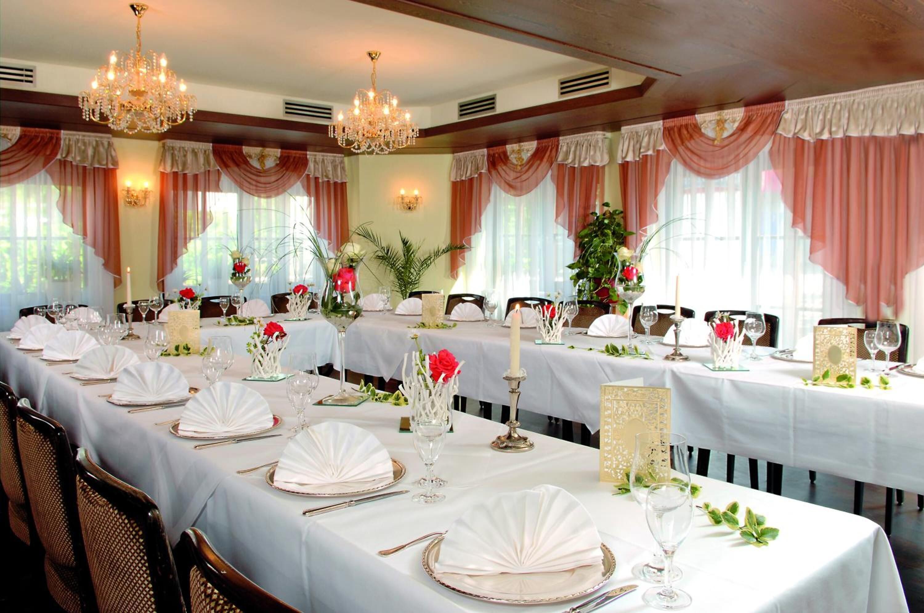 A banquet hall with long tables set for a formal event, featuring chandeliers, draped curtains, and floral centerpieces.