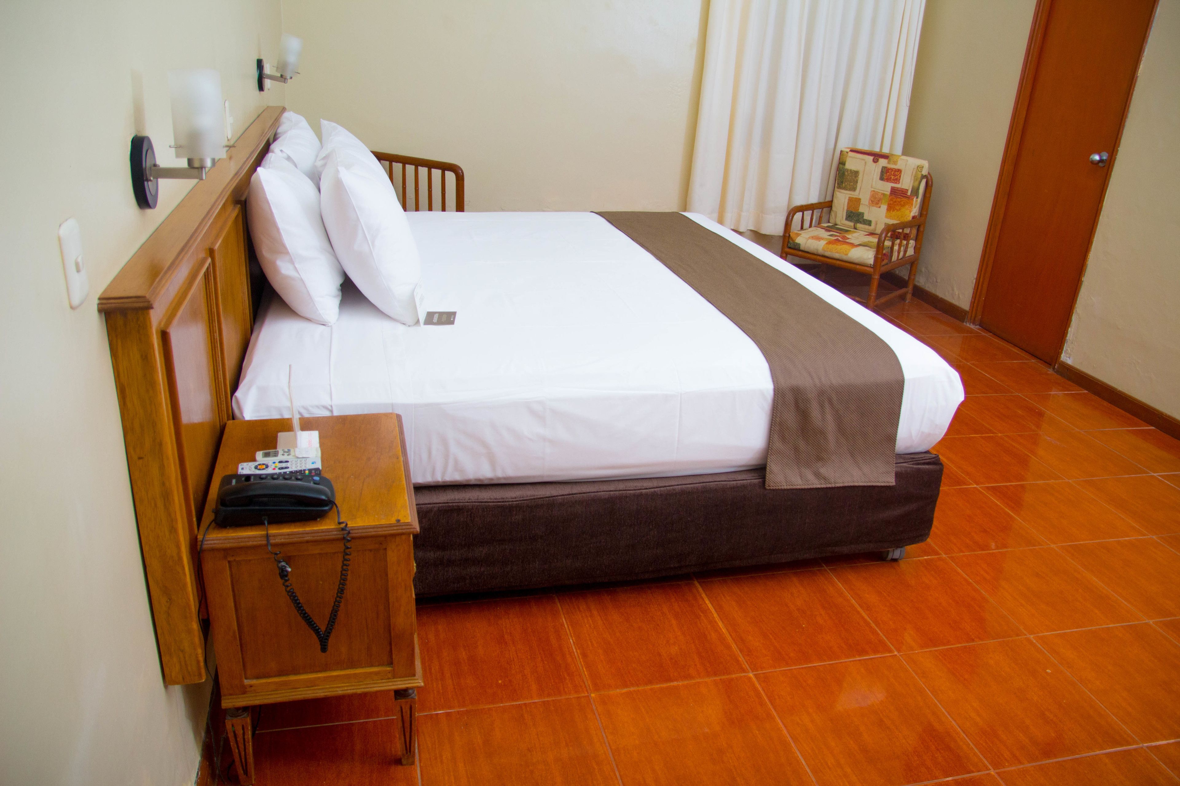 Una cama bien hecha con cabecera de madera, una mesita de noche con un teléfono y una lámpara, y una silla con asiento acolchado.