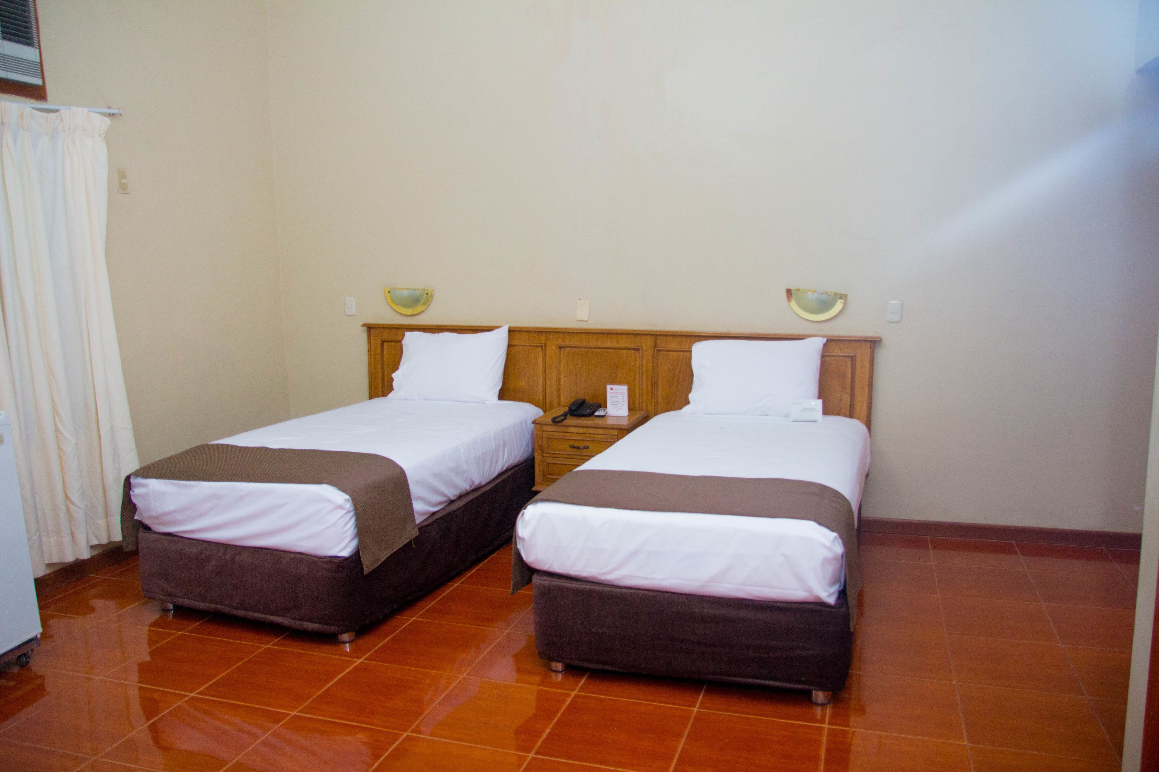 Habitación de hotel con dos camas, cabecero de madera, mesita de noche pequeña con lámpara y suelo de baldosas.