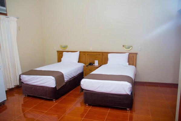 A hotel room with two beds, a wooden headboard, a small bedside table with a lamp, and a tiled floor.