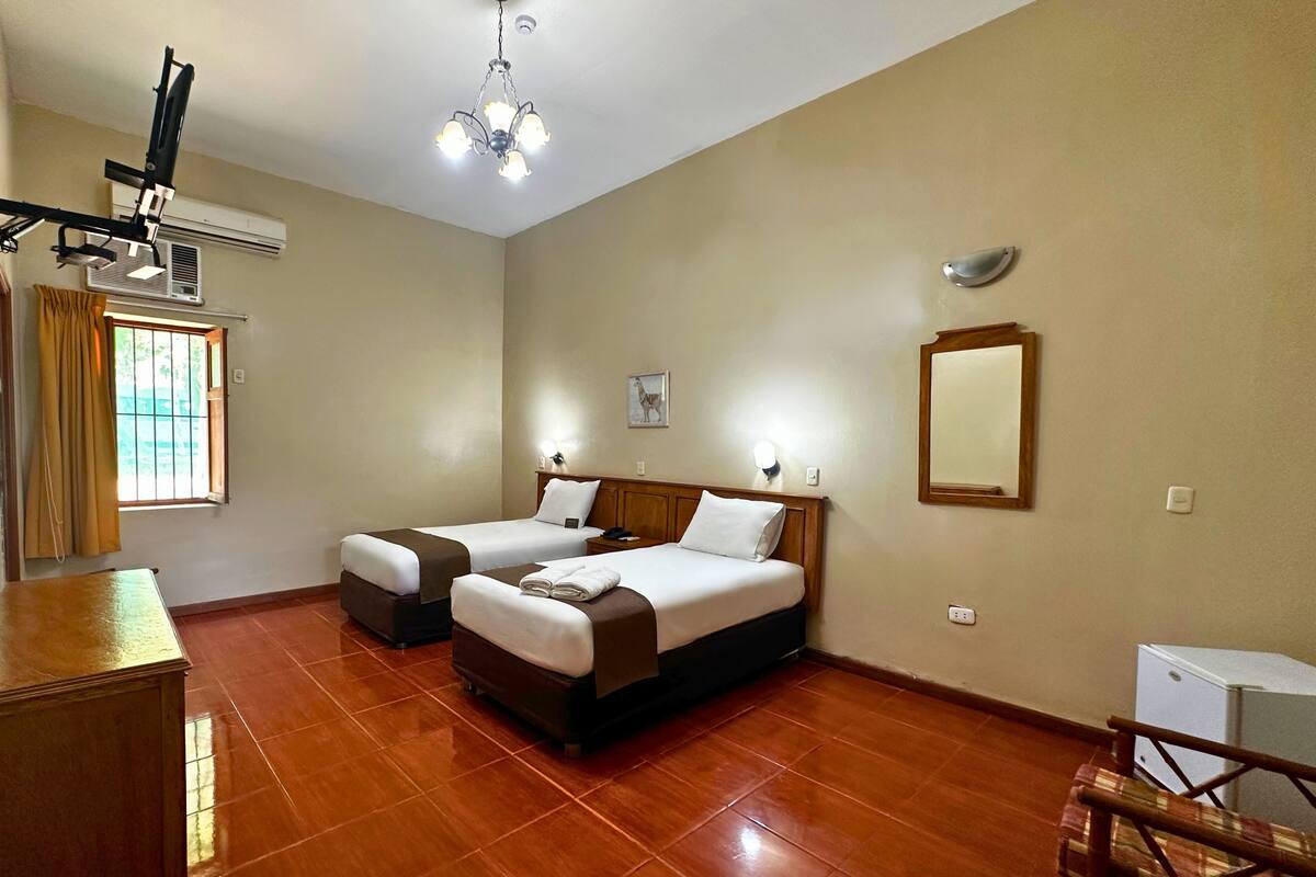 A hotel room with two beds, a desk, a chair, a ceiling fan, an air conditioning unit, a window with curtains, a wall-mounted mirror, and a small refrigerator.