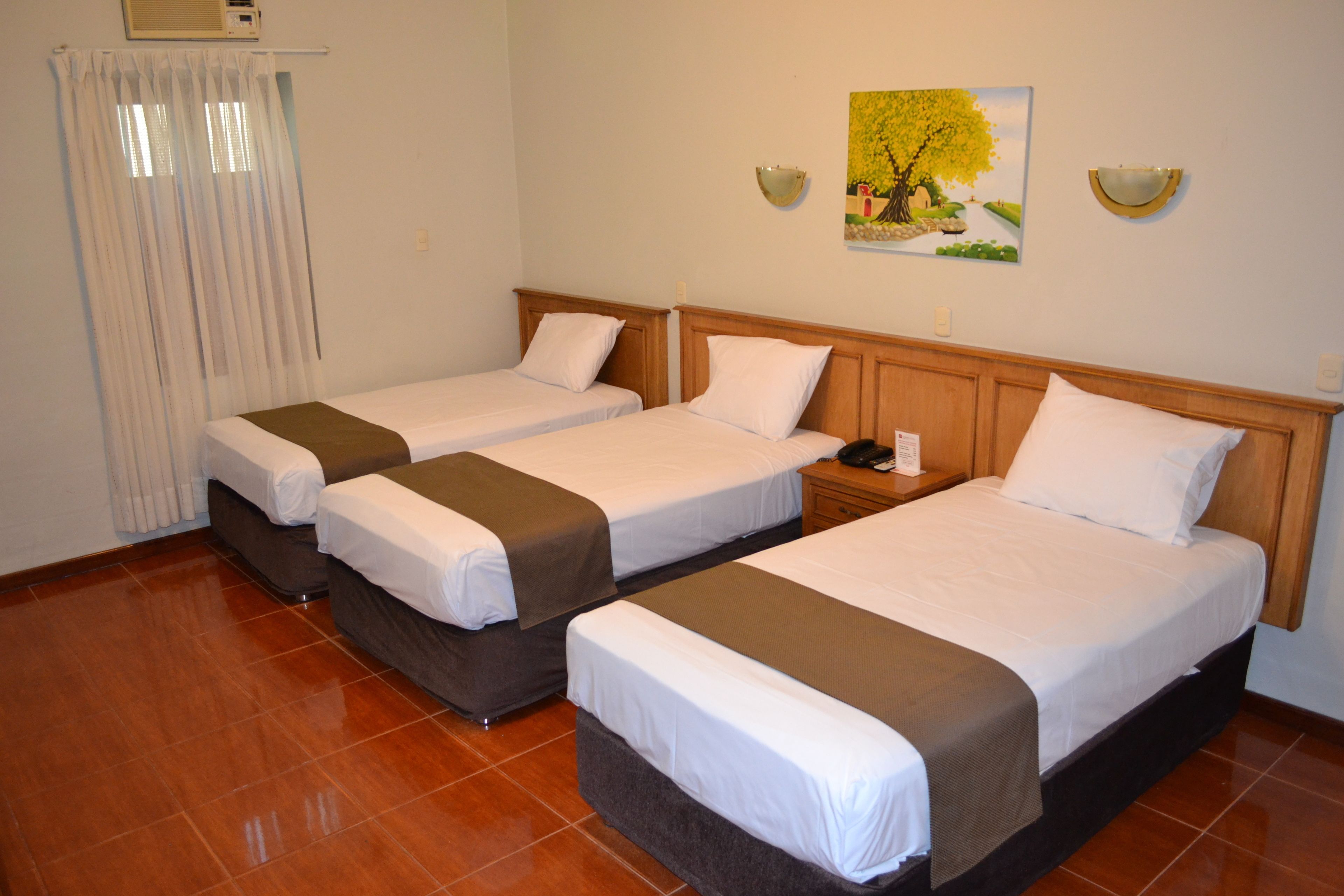 A hotel room with three single beds, a wooden headboard, a nightstand, a wall-mounted lamp, and a painting of a tree.