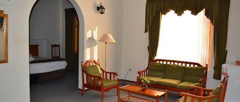 A hotel room with a bed, a sofa, two armchairs, a coffee table, a lamp, and a window with curtains.