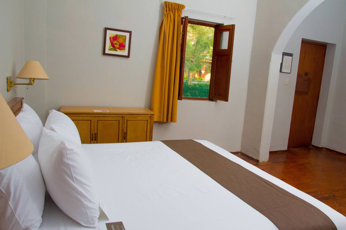 A hotel room with a bed, a nightstand, a lamp, a window with curtains, a door, and a framed picture on the wall.