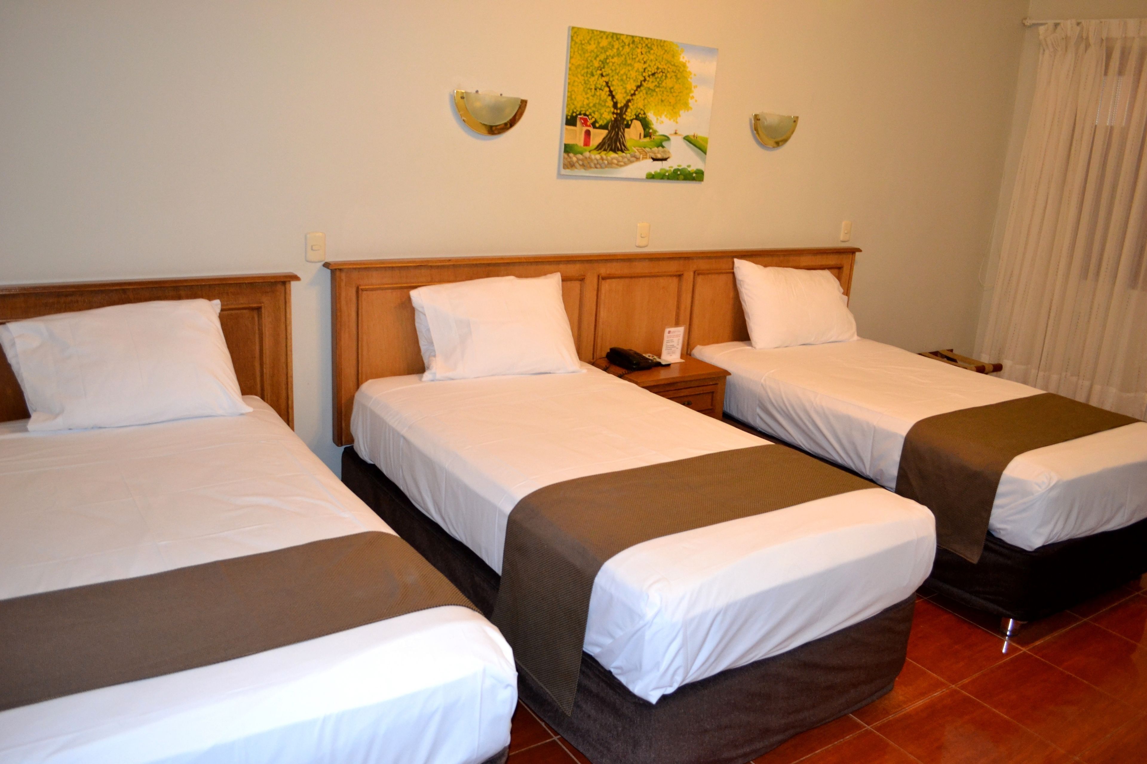 A hotel room with three single beds, each with a wooden headboard and white bedding. There is a painting of a tree on the wall above the beds.