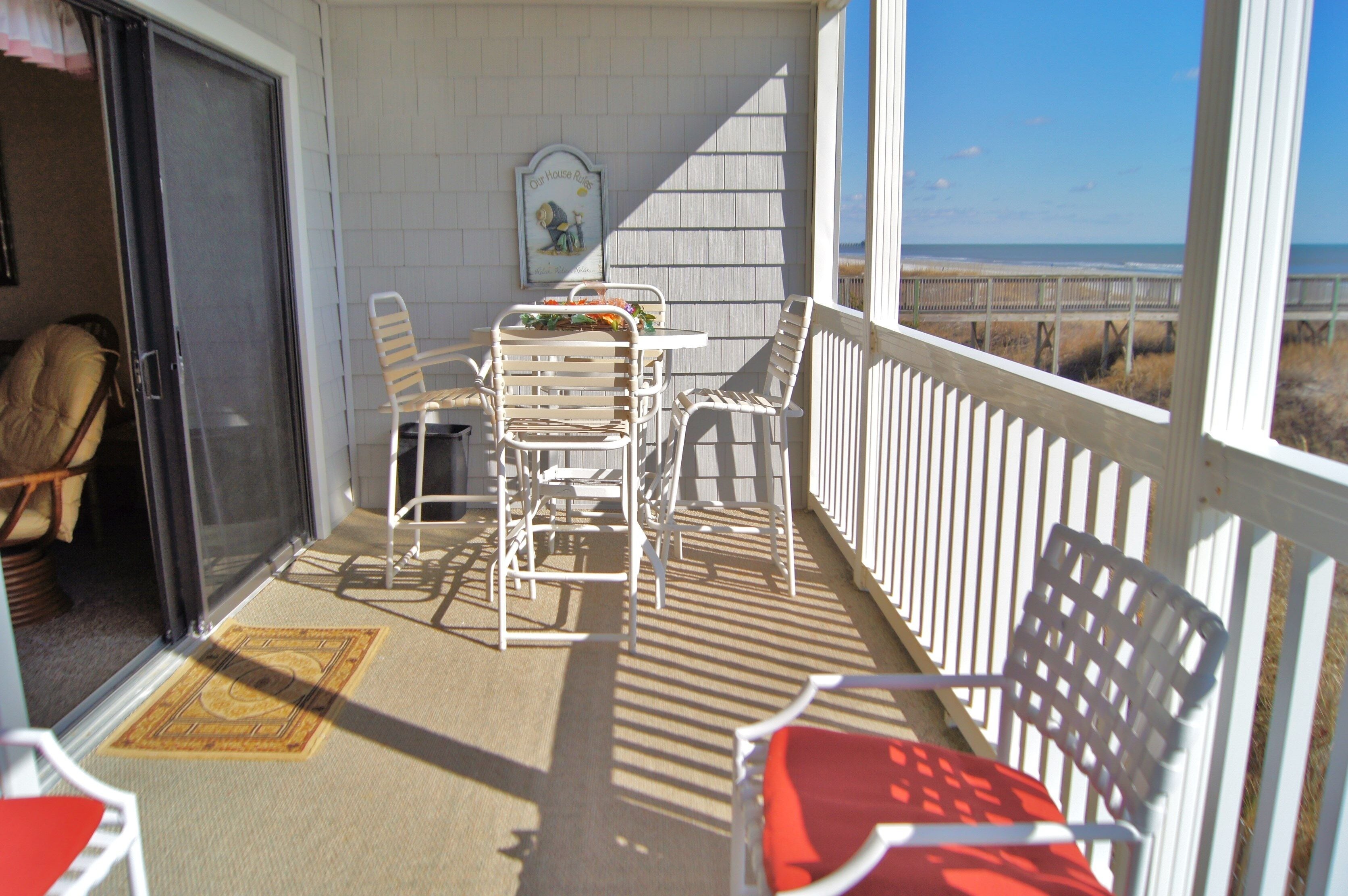 A balcony with white chairs and a table, a door leading to a room, and a view of the beach.