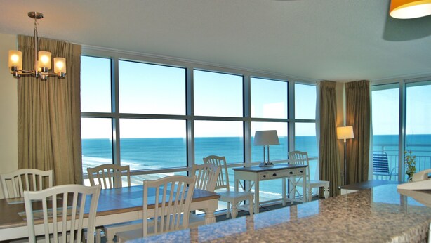 A room with a large window overlooking the ocean, a dining area with white chairs and a table, and a small desk with a lamp. - Seaside Resort 1003 3 Bedroom Condo by RedAwning (North Myrtle Beach)