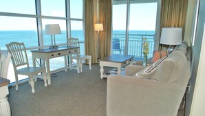 A hotel room with a large window overlooking the ocean, a white sofa, a small table with a lamp, and a chair. - Seaside Resort 1003 3 Bedroom Condo by RedAwning (North Myrtle Beach)