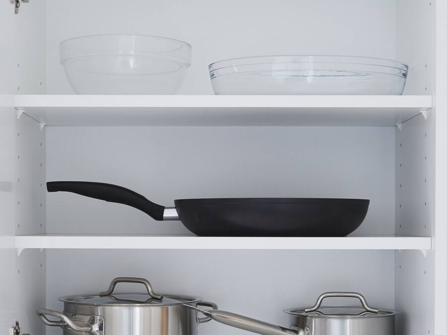 A kitchen cabinet with a clear glass bowl, a black frying pan, and two stainless steel pots.