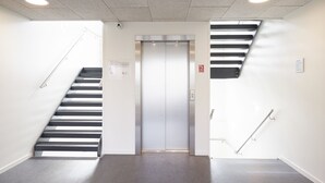 Ein moderner Aufzug mit Metalloptik, eine Treppe mit schwarz-weißen Stufen und ein grauer Boden.