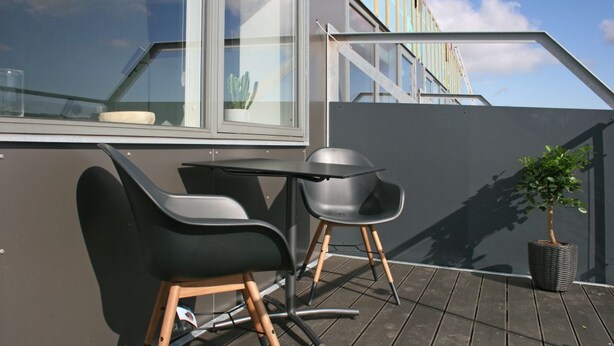 Eine Dachterrasse mit Tisch und zwei Stühlen, einem Blumentopf und Blick auf ein Gebäude im Hintergrund.