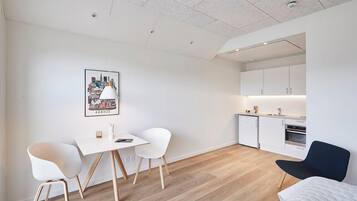 A modern, minimalist room with a wooden floor, white walls, a dining table with two chairs, a bed, and a kitchenette.