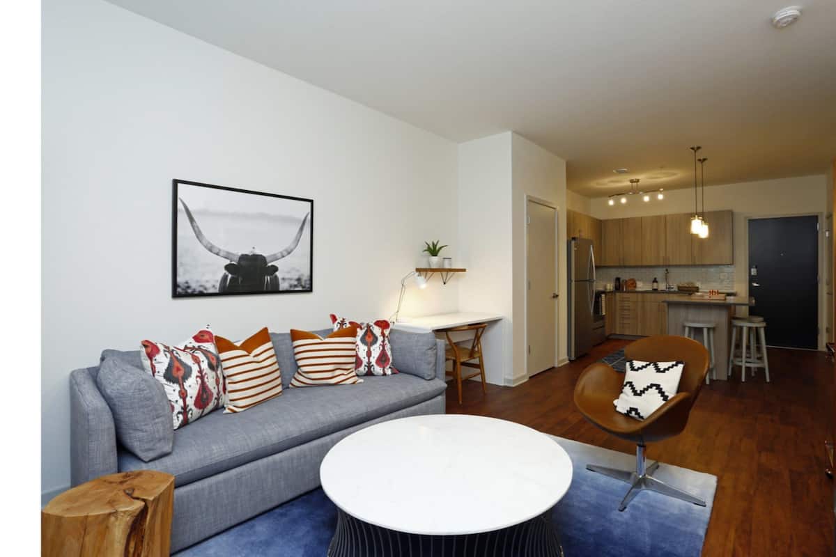 Ein modernes Wohnzimmer mit einem grauen Sofa, einem runden Couchtisch und einem braunen Sessel. Im Hintergrund befindet sich eine Küche mit Holzschränken und einer Pendelleuchte.