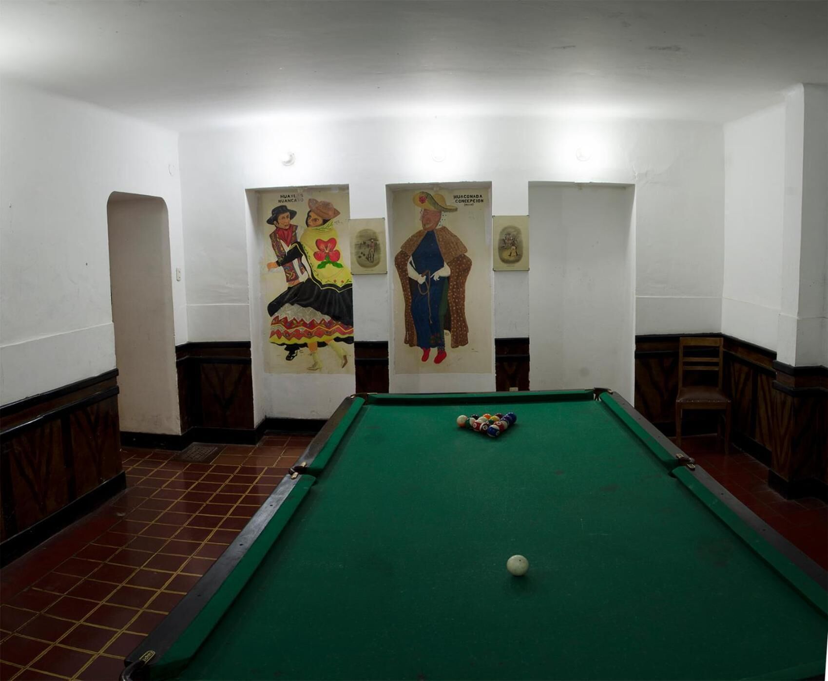A pool table with balls in a room with white walls and tiled flooring. There are three framed pictures on the wall.