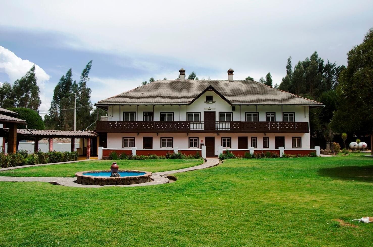 A two-story building with a tiled roof, a central balcony, and a small fountain in the front yard.