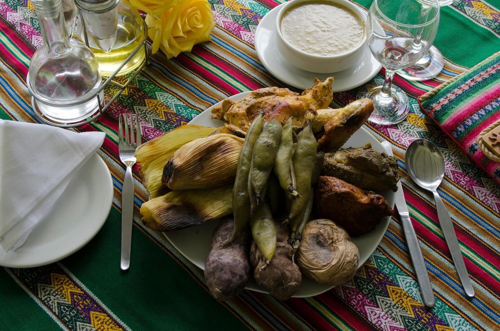 A plate of food with corn, potatoes, and a roasted chicken leg, accompanied by a bowl of soup and a glass of wine on a colorful tablecloth.