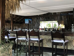 A bar area with wooden chairs and a stone counter, featuring a thatched roof and a poster on the wall.