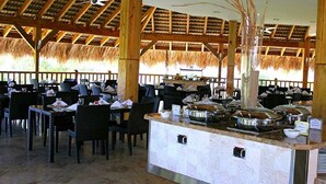 A buffet-style restaurant with multiple dining tables and chairs, a central serving counter with food containers, and a thatched roof structure. - Lifestyle Kosher Cofresi Suites (Puerto Plata)